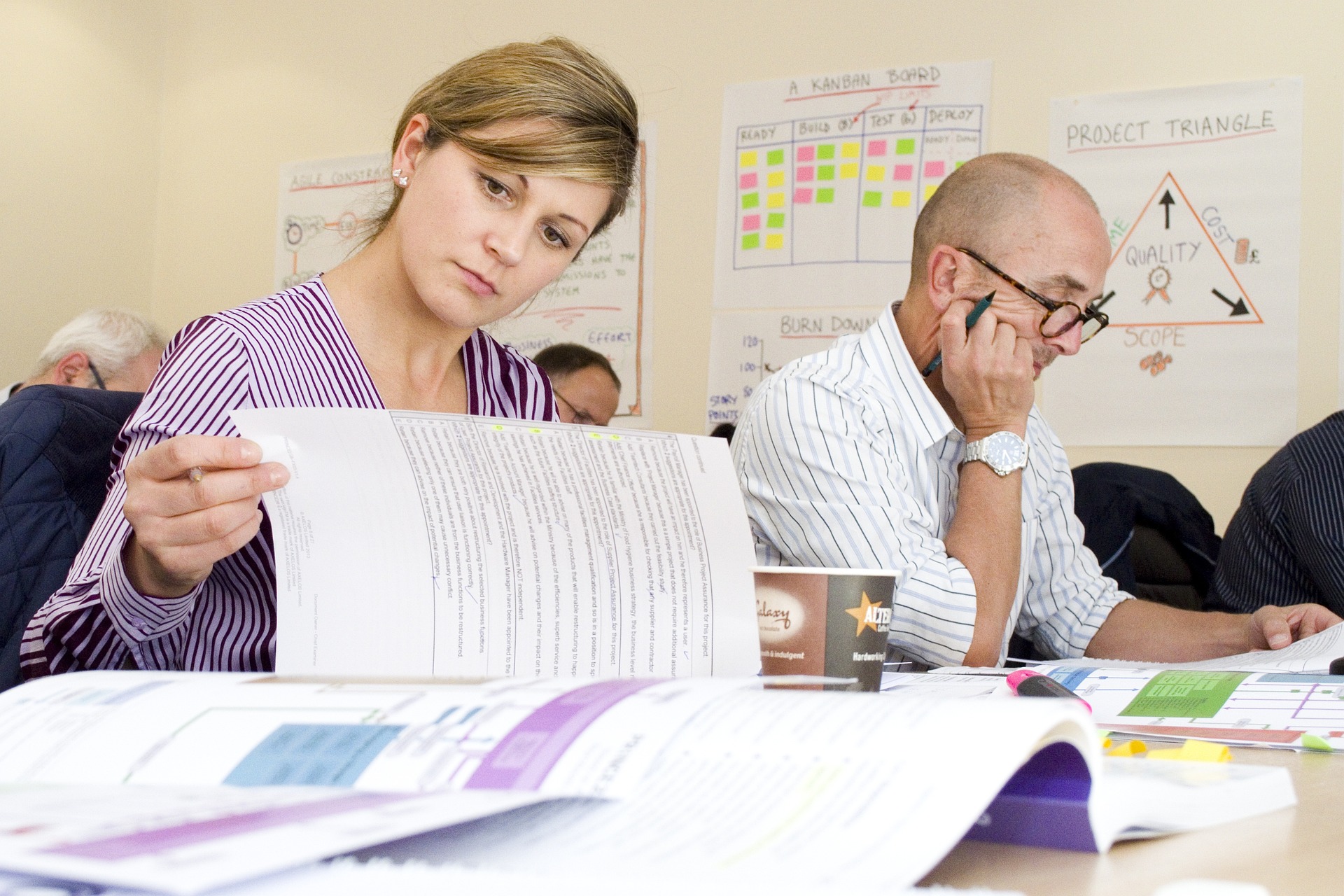 Two project managers sit at a table leafing through documents with posters behind them showing project process